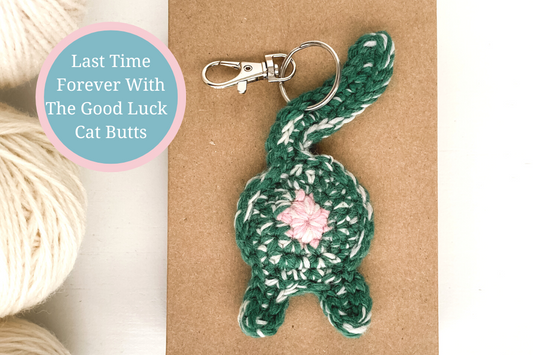 Green cat butt keychain on a Kraft box next to white yarn with the text Last Time Forever With The Good Luck Cat Butts in a teal circle