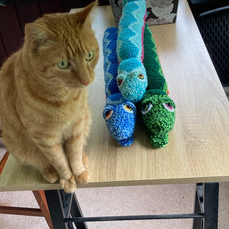 Orange cat next to two small snake dolls and one large snake dolls, all  on a wooden table.