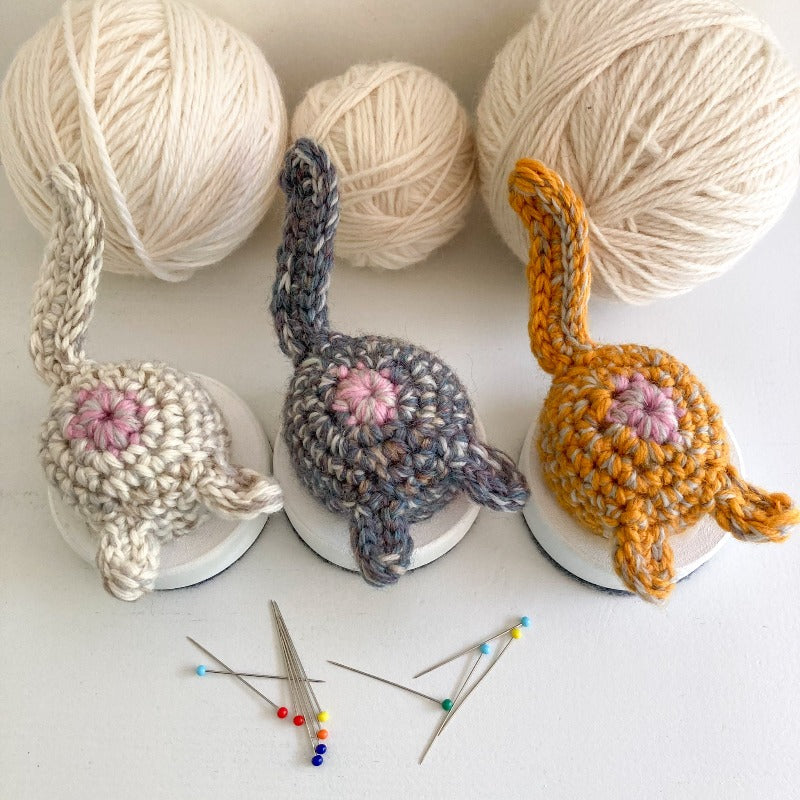 Crocheted white, gray purple, and orange cat butt pincushions in front of 3 balls of white yarn and behind some pins