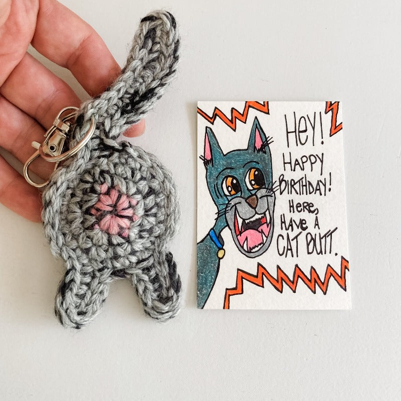 gray cat butt keychain with hand for scale and one of a kind birthday art card