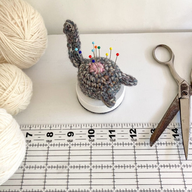 crocheted gray purple cat butt pincushions with pins sticking out of it. It's next to silver scissors, 3 balls of white yarn and a clear sewing ruler.