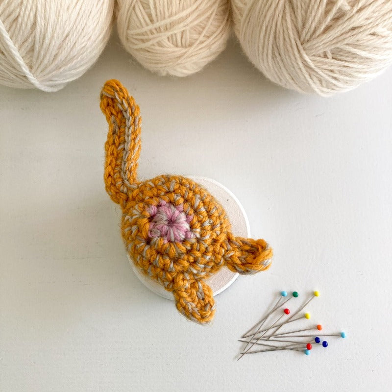 crocheted orange cat butt pincushion with 3 balls of white yarn above it and a small pile of pins underneath it.